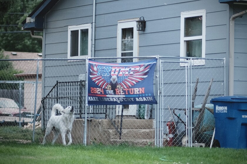 A Trump 2024 campaign flag hangs on a chain-link fence outside a house in Dickinson, North Dakota. The flag, displaying an eagle with wings spread, reads “Trump 2024 The Return” and “Make Liberals Cry Again.” A white dog stands nearby, barking.