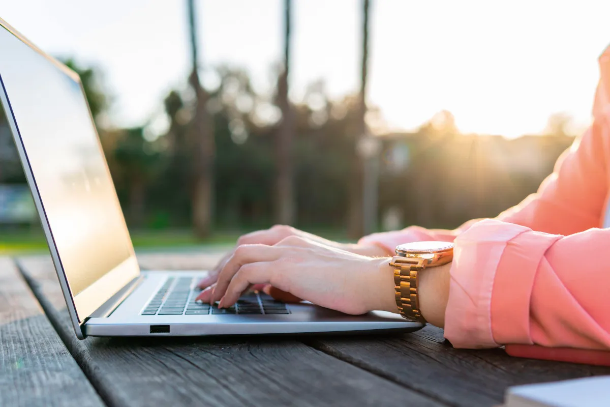 A person typing on a laptop on top of a wooden table outside.