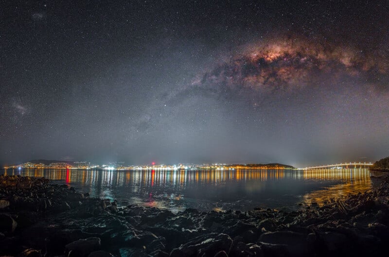 A star-filled night sky with the Milky Way galaxy arching overhead reflects on a tranquil body of water. Distant city lights line the horizon, casting vibrant reflections on the surface. Rocky shore in the foreground.