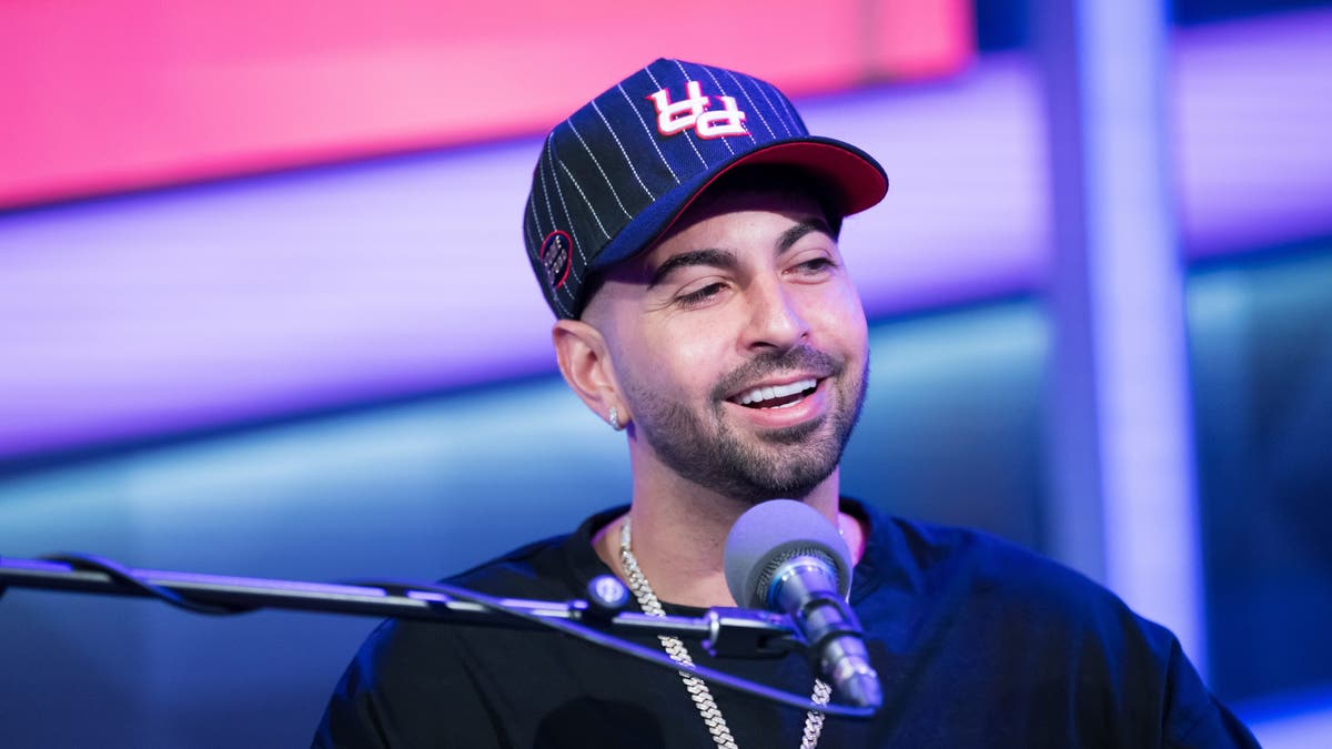 man sitting behind podcast microphone, smiling with black baseball cap on