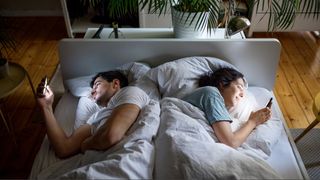 A couple laying in bed with white sheets with their backs to each other as they both scroll on their phones.