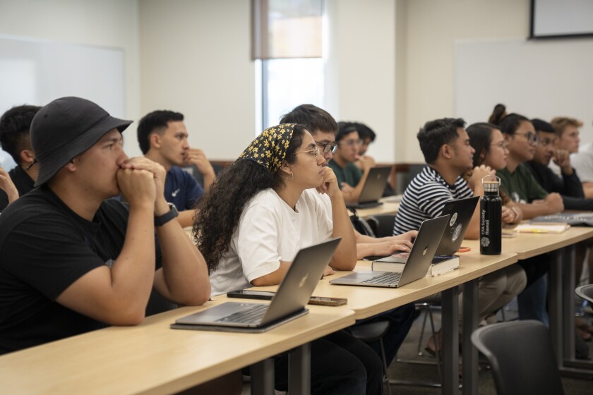 Students sit at tables with their laptops open while looking towards the front.