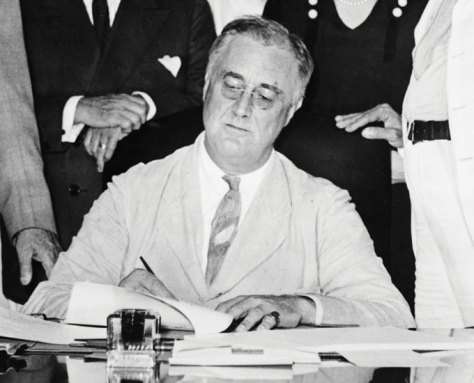 A black-and-white photo of President Franklin Roosevelt sitting at a desk and signing papers