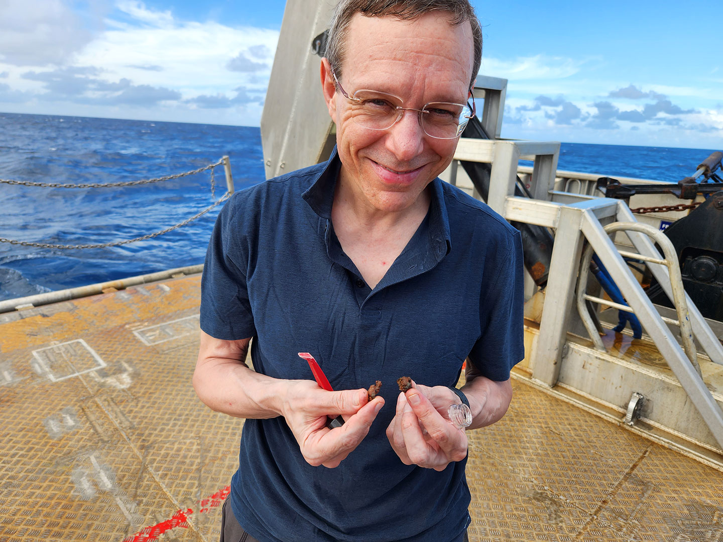 a photo of Avi Loeb, a smiling older man standing on a boat, holding two small bits of space debris in his hands