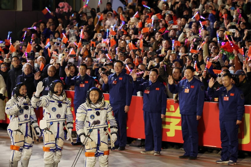 three chinese astronauts dressed in their white spacesuits walks in front of an adorning crowd of chinese people holding flags