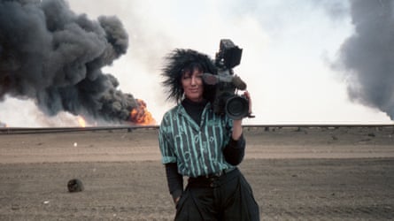 She holds a camera in the desert, smiling, while a fireball blooms behind her