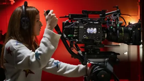 Netflix Anna Kendrick on the set of Woman of the Hour. She is wearing headphones and operating a large film camera. 