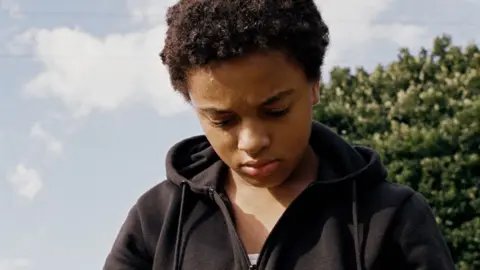 Mubi Nykiya Adams as Bailey in Bird, pictured in a black hoodie and looking at the ground