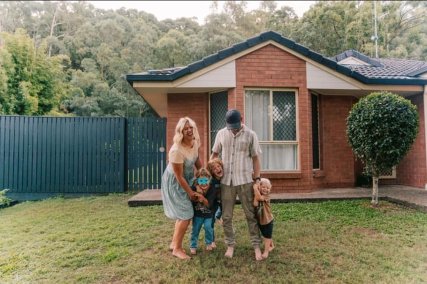 A young man and woman smiling in front of a house, with three young kids by their sides.