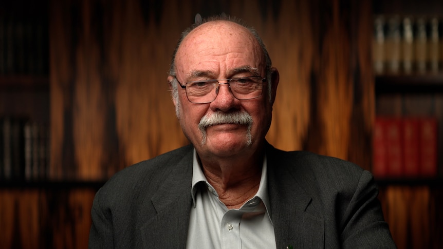 Older man with grey moutache wearing a suit looking down the barrel of the camera. 