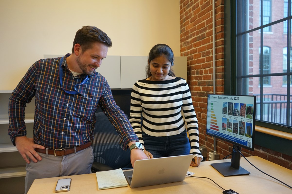 A man in business attire smiles while pointing to a laptop screen while standing next to a young woman in an office.