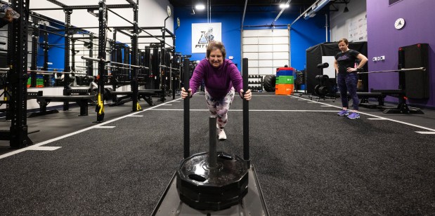 A woman pushes a weighted sled while her trainer watches