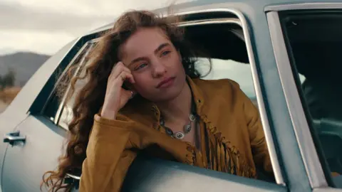 Netflix Autumn Best as Amy in Woman of the Hour. She is leaning out of the passenger side of the car and is wearing a mustard coloured suede jacket with tassels on. 