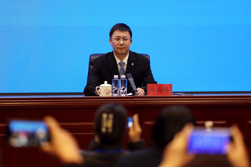 a chinese man sits behind a desk conducting a press conference speaking on behalf of the China Manned Space Program