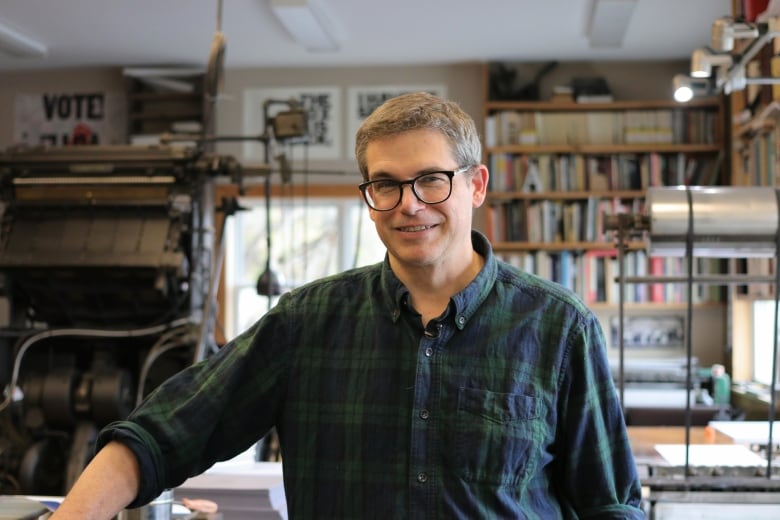 A person with short hair wearing a flannel shirt and glasses smiles in a workshop with many printing machines and books aound.