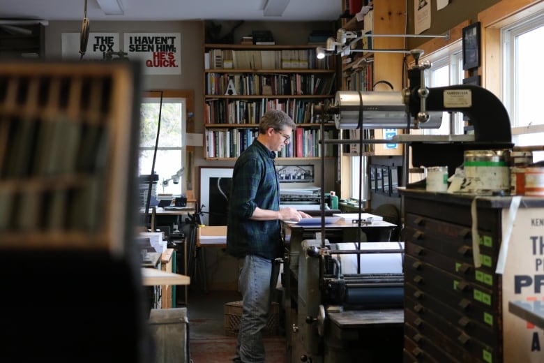 A person in a flannel shirt and jeans operates a press in a workshop with many things around.