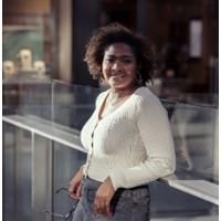 Photo of Ane Ogbe standing against a glass screen. Ane is wearing a white cardigan and holding her glasses.