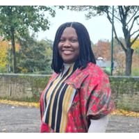 Photo of Blessing Airhihen. Blessing is standing outside near a brick wall and some trees. She is wearing a red, yellow and black top.