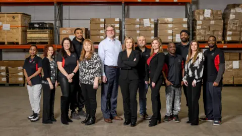 Lucerne International Mary Buchzeiger (centre) stands in a row with her team at a Lucerne International warehouse. The team look at the camera, with carboard boxes piled up on shelves behind them.