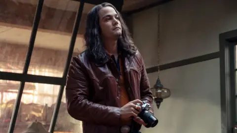 Netflix Daniel Zovatto as Rodney Alcala in Woman of the Hour. He has long hair, a burgundy leather jacket and is holding a camera.