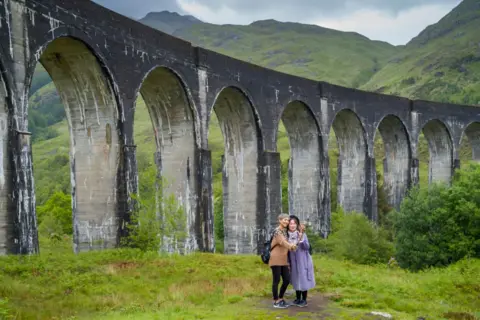 Getty Images Two tourists stand in front of Glenfinnan Viaduct, a dramatic railway arch in the West Highlands in Scotland. There are taking a selfie with a selfie stick