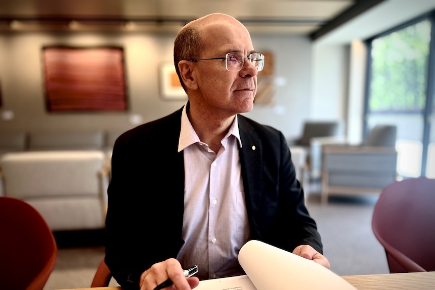 An older man wearing a business shirt, suit jacket and glasses sits at a desk looking into the distance.