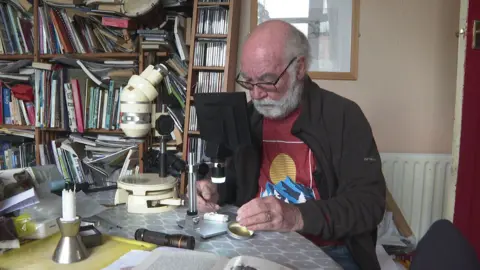 Paul Moore examining flies under a microscope with overflowing bookcases behind him