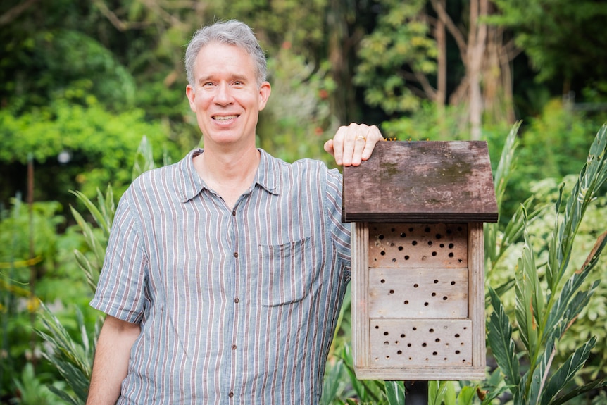 A man leans on a beehive and smiles at the camera