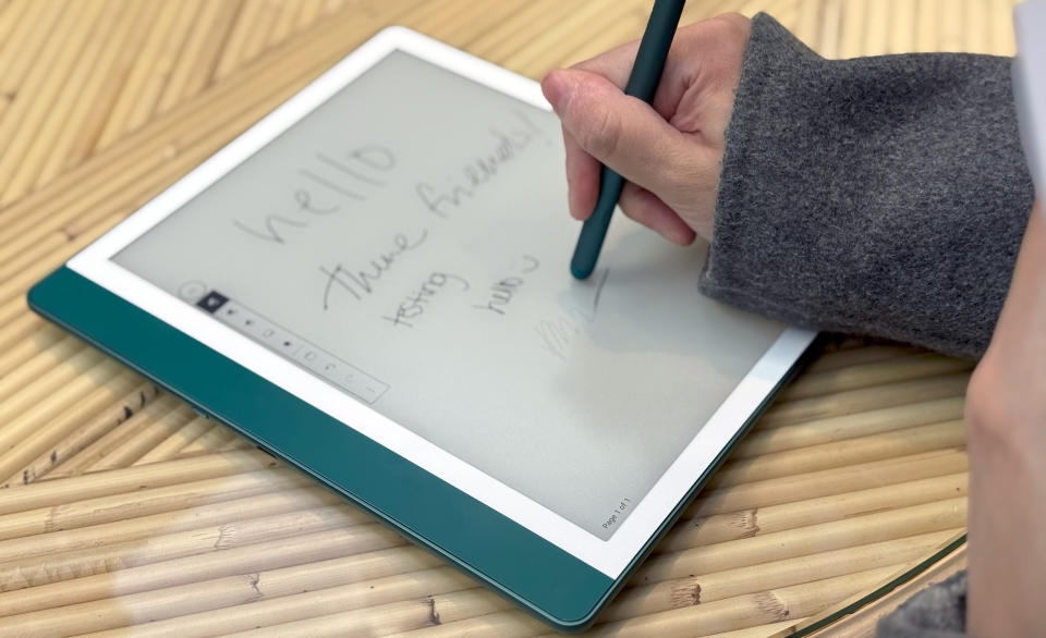 The Amazon Kindle Scribe on a table, with a hand holding a pen to its screen, erasing some words.