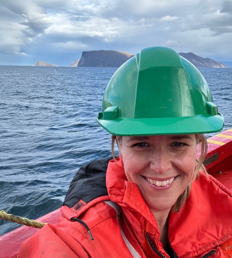 Woman in green hard hat and red jacket smiling while leaning against the edge of a boat. Behind her is water with a mountain in the background.