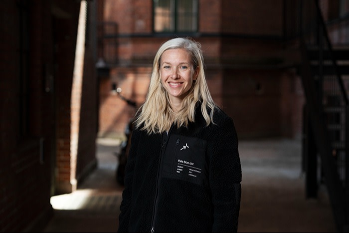 A woman with long blonde hair, wearing a black fleece jacket, stands smiling outdoors in a narrow alley with red brick buildings