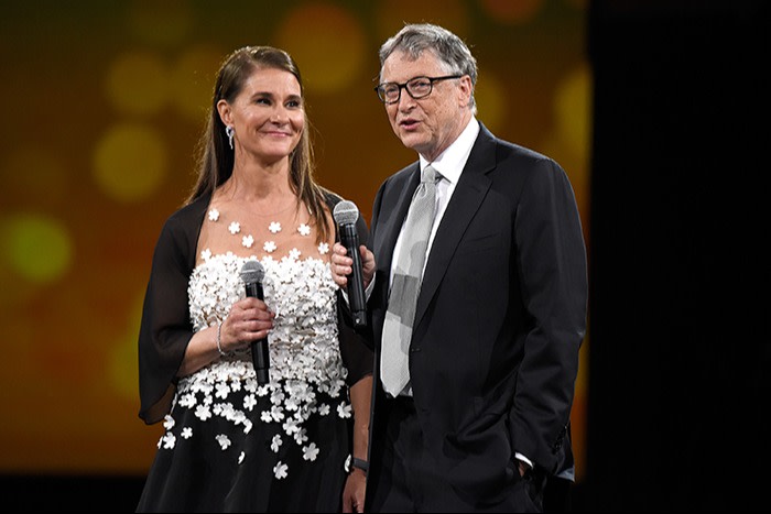 A woman in a black dress with white flowers and a man in a black suit hold microphones during a formal event on stage