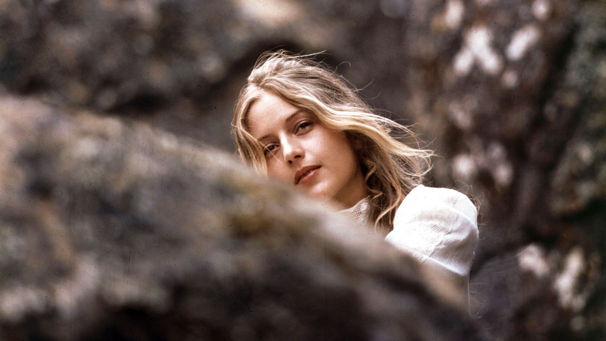 A young girl in a white dress sits between rocks in a still from 