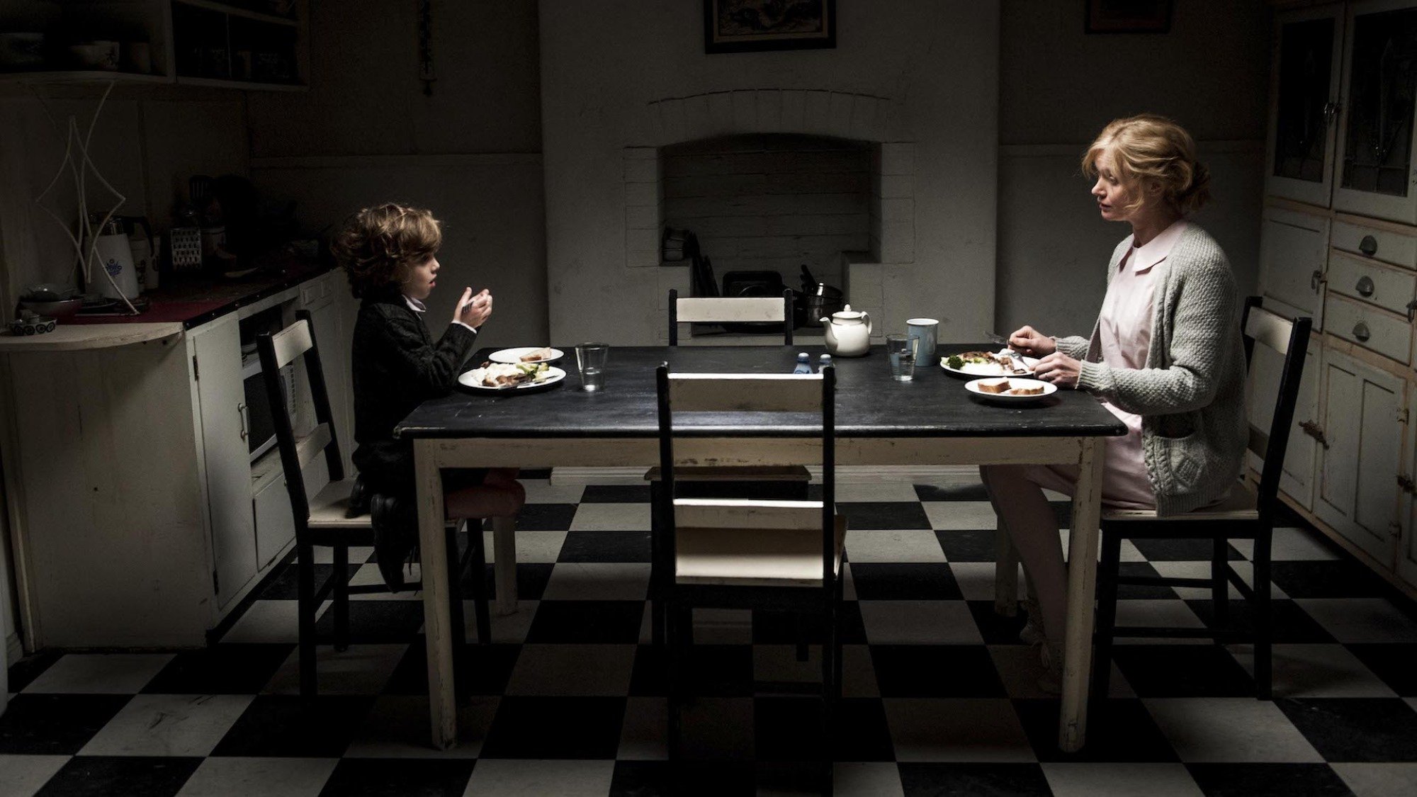 A child and a woman sit at opposite ends of a kitchen table in low lighting in 