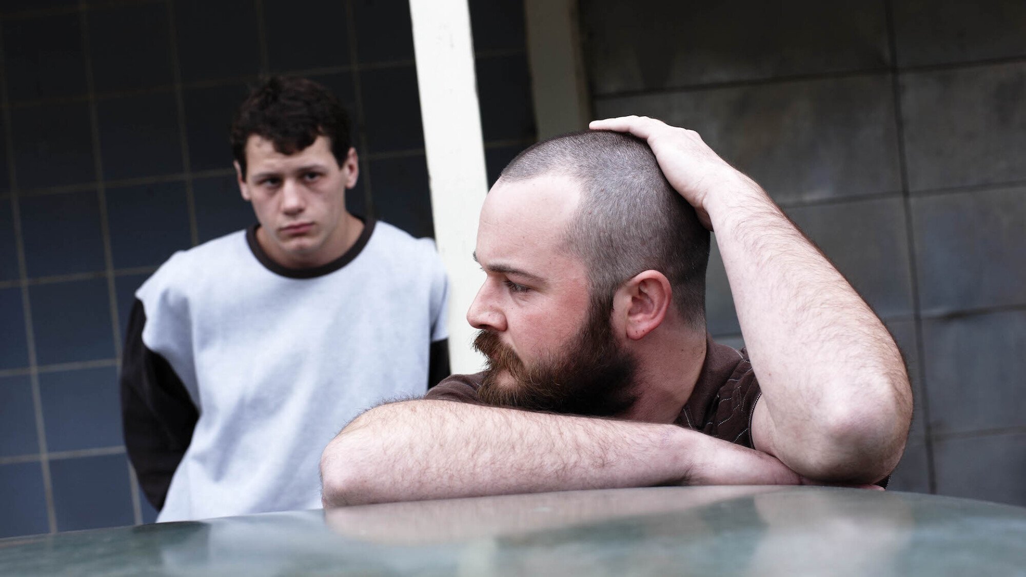 Two men stand looking grim beside a car in the film 