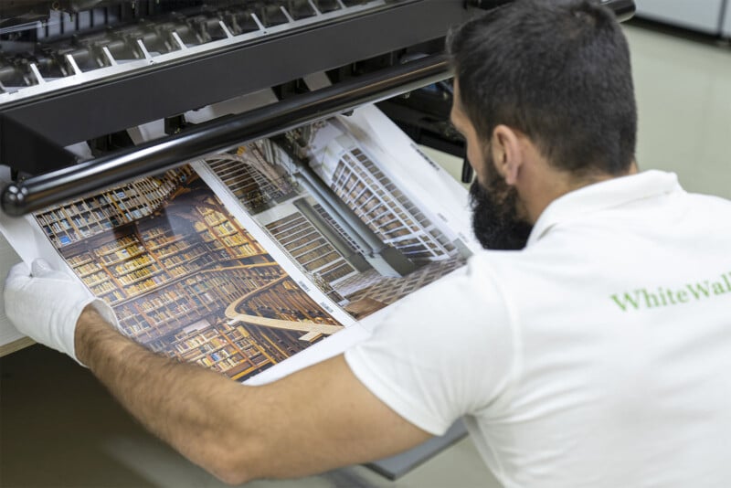 A man in a white shirt examines two large photographs of library interiors being printed. The images show shelves filled with books and architectural designs. He is wearing white gloves and carefully checks the quality of the prints.