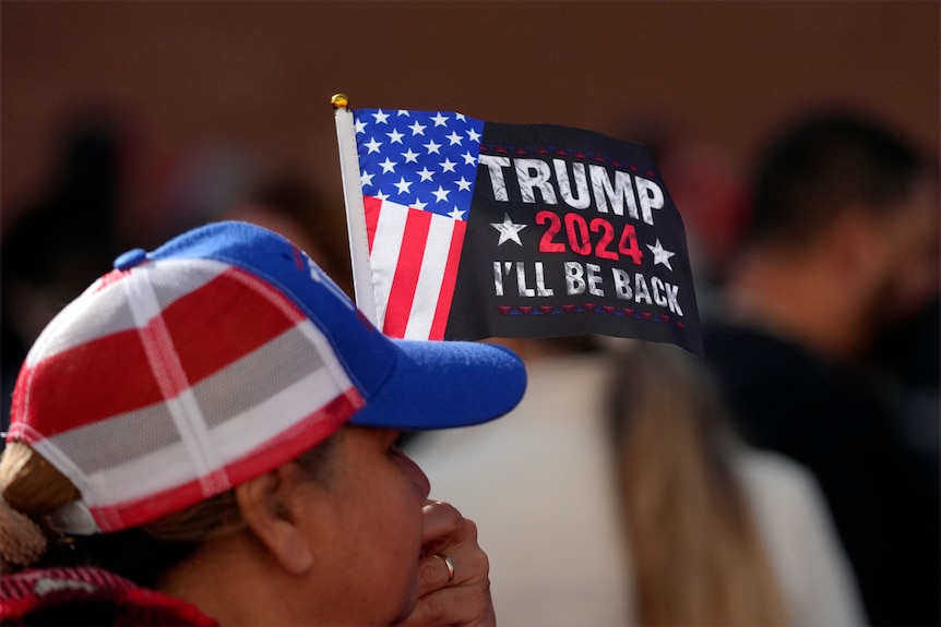 A Trump supporter wearing a cap holds a sign that says Trump 2024 I'll be back.