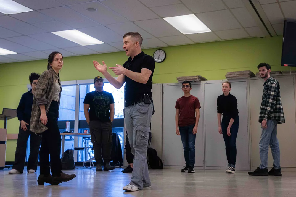 Sebastian Molnar in a dance studio with a green wall talking to six of his students that surround him.