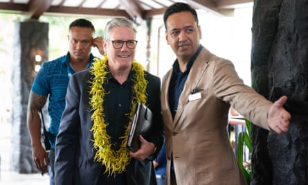 A man with his arm held out stands to the side as he directs Starmer forward. The PM is wearing a yellow floral garland