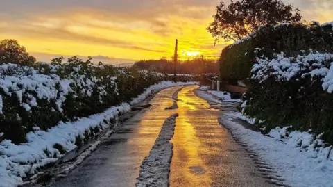 arron | BBC Weather Watchers Sunset on a country road. the sky is golden and reflecting onto the road surface which has a strip of snow in the middle where car tyres have not melted the ice.  the hedgerows on either side of the road are also covered in snow. 