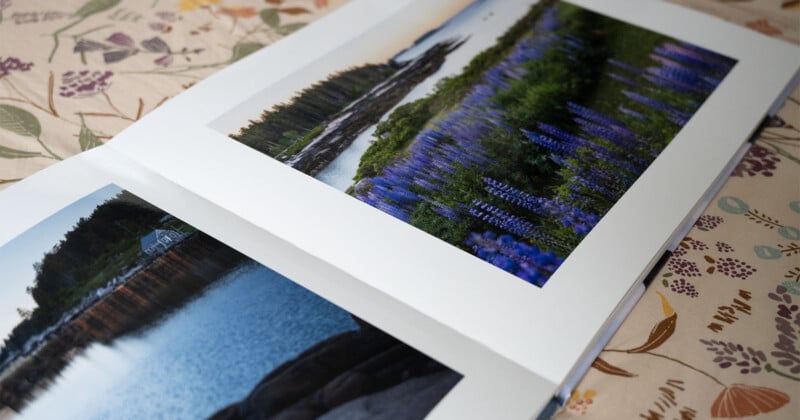 An open photo album displays two scenic images. The left page shows a calm lake with a reflection of a house, while the right page features a field of purple flowers by a river. The album rests on a floral-patterned tablecloth.