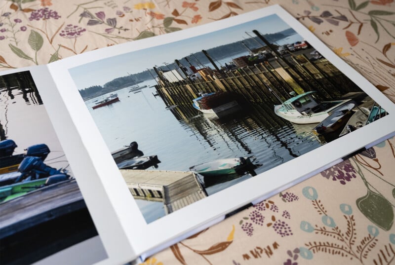 An open book displaying a photograph of a serene harbor, featuring boats moored at wooden docks under a clear blue sky. The image is set against a floral-patterned tablecloth.