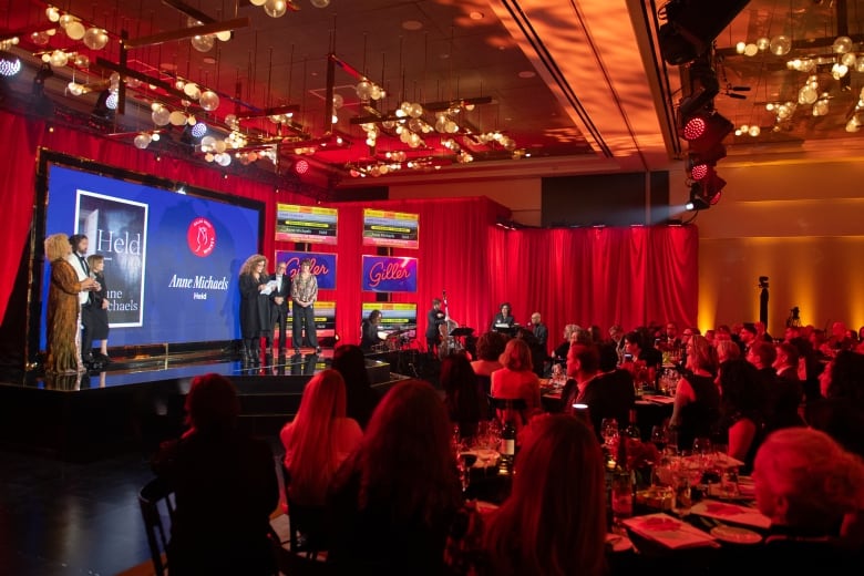 A photo of a ballroom with a tables facing a stage with a screen behind it. A woman wearing a black suit with curly hair stands in the middle of the stage.