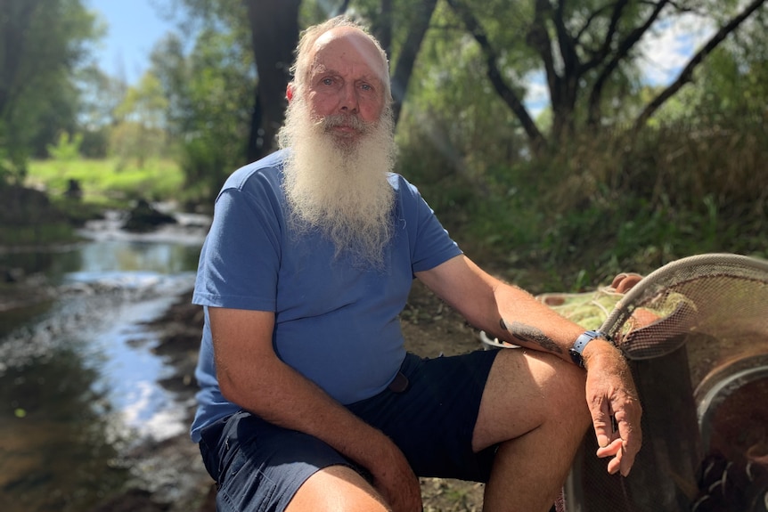 A man with a grey beard sits next to a small stream.