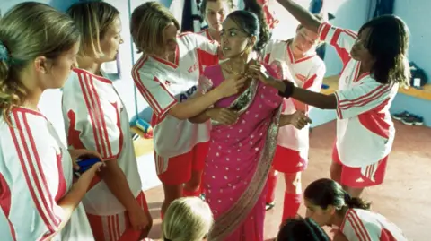 Getty Images A scene from Bend it Like Beckham in which a group of white girls in football kit fit a pink sari with gold embroidery on to an Indian teenage girl, Parminder Nagra