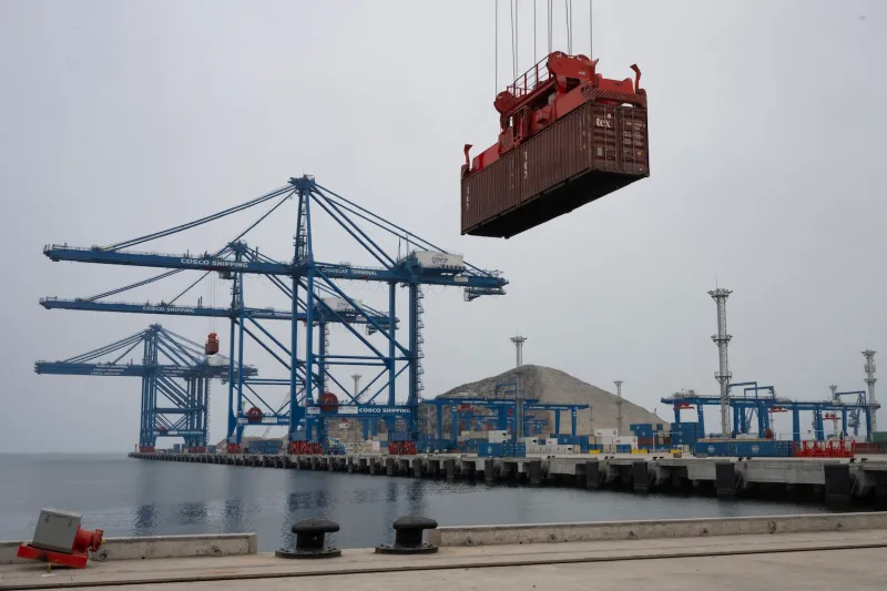 A long dock stretches into the water at a port in Peru. Massive blue cranes are perched along its length, and one behind the camera has lifted a metal shipping container, so it hangs at the top of the image against a pale gray sky.