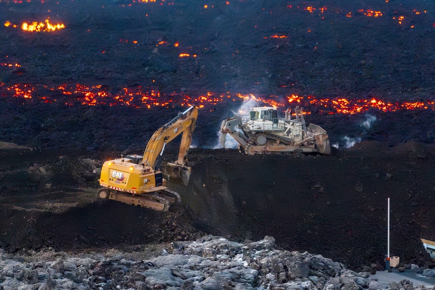 Excavators dig out a large ditch as lava glows in the background