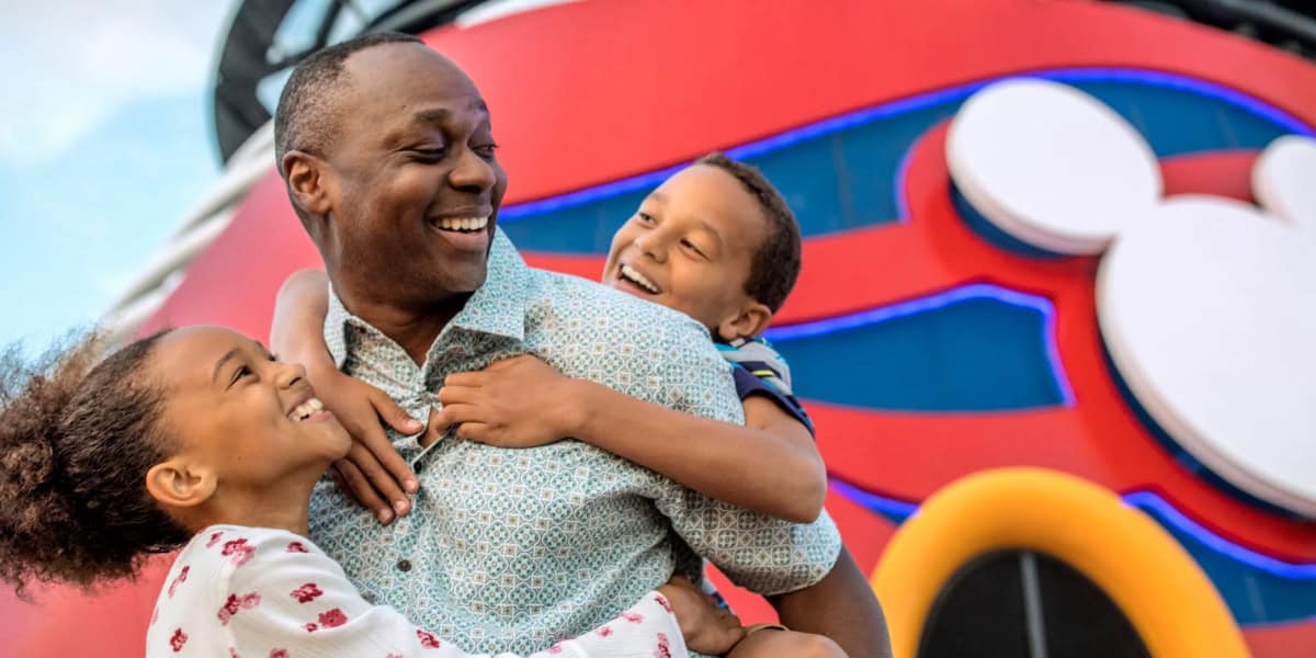 A joyful man embraces two smiling children, one girl and one boy, in front of a vibrantly colored structure featuring the iconic Mickey Mouse logo. The backdrop includes bright red and blue elements, suggesting a fun, themed setting for this Disney destination.