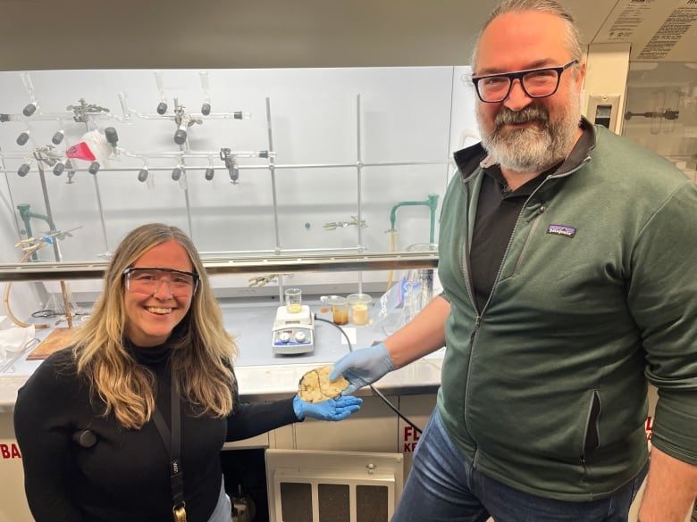 Woman with blond hair in black sweater and man standing with green sweater and he's passing her a off-yellow blob. Behind them are scientific equipment and they are wearing blue gloves.