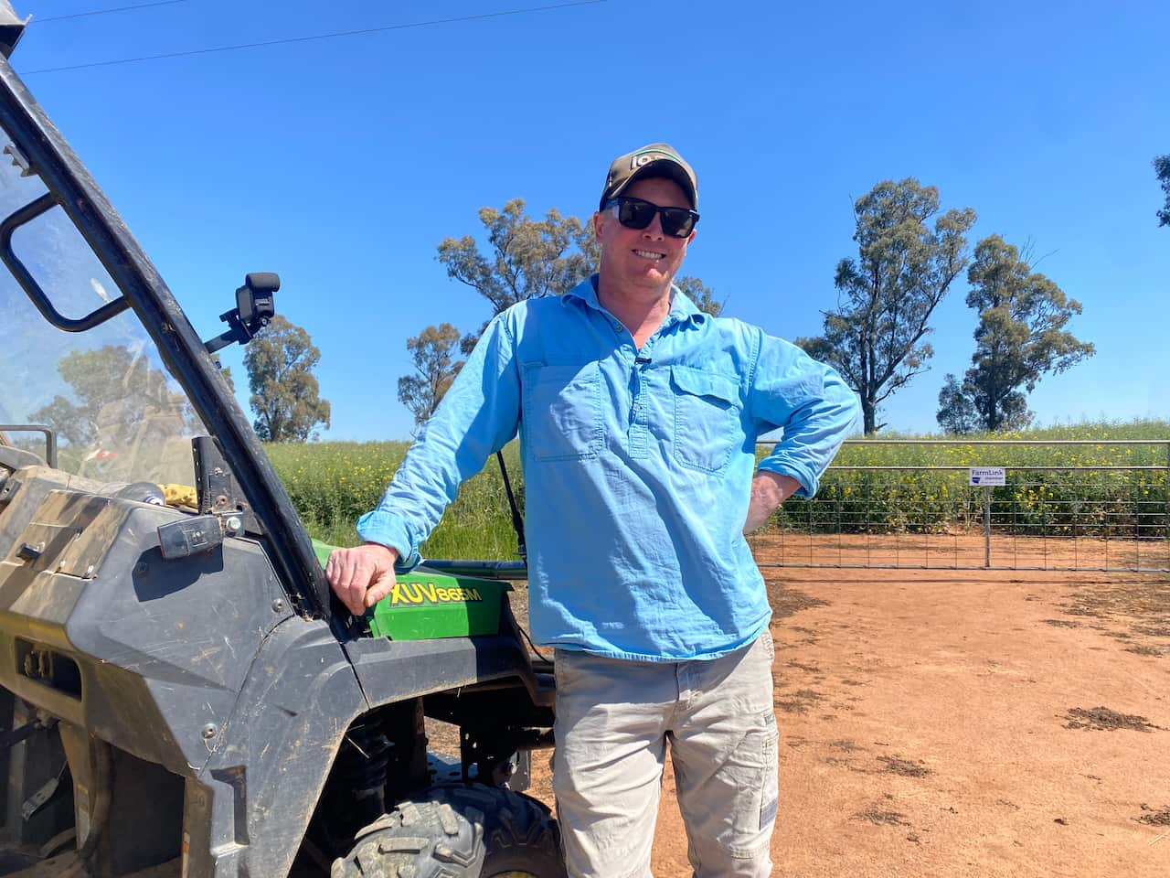 A man wearing a blue shirt, trousers and a cap smiles while leaning on a tractor. 